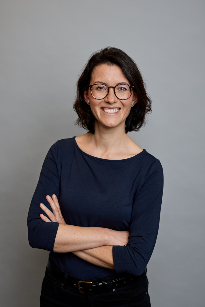 Lisett mit verschränkten Armen. Sie trägt ein dunkelblaues Shirt mit Dreiviertel-Ärmeln und lächelt herzlich.