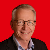 Das Bild zeigt einen Mann mit grauen Haaren, Brille, glatt rasiert und einem leichten Lächeln 