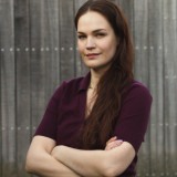 Die Kandidierenden schaut freundlich und gleichzeitig, hoffnungsvoll und bestimmt in die Kamera. Ihre Arme sind verschränkt. Sie hat lange rotbraune Haare. 