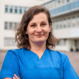 Ingrid Petri Brünette Krankenschwester im mittleren Alter steht lächelnd und armeverschränkend vor einem Krankenhaus.