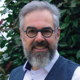 Portrait von Michael Kefer, graue Haare, Bart- und Brillenträger. Im Hintergrund eine grüne Hecke