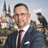 Daniel Wald im dunkelblauen Anzug mit weißem Hemd und bordeauxfarbener Krawatte. Im Hintergrund sind der Merseburger Dom und Schloss, sowie eine Rabenstatue aus der Rabensage, mit dem Ring zu sehen.