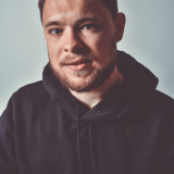 Moritz Riedacher Profilbild von Moritz Riedacher. Es zeigt ihn sitzend, mit geraden Blick in die Richtung des/der Betrachter:in blickend. Er trägt einen schwarzen Kapuzenpullover, hat einen Bart und kurze Haare.