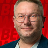 Alternativtext für das Bild: Porträt eines Mannes mit Brille und kurzem, blond-grauem Haar, der freundlich und selbstbewusst in die Kamera blickt. Er trägt ein dunkles Sakko über einem hellen T-Shirt. Im Hintergrund ist eine rote Fläche mit grafischen SPD-Schriftzügen zu sehen.