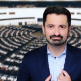 Ein Mann Mitte 40 mit einem Blauen Anzug. Im Hintergrund ist der Plenarsaal des EU-Parlaments