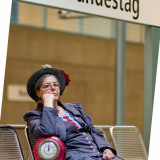 Andrea Schulteisz sitzt auf einer Bank im U-Bahnhof Bundestag. Neben ihr steht eine Tasche mit einer großen Uhr vorne drauf. Die Zeiger stehen auf kurz vor 12. 