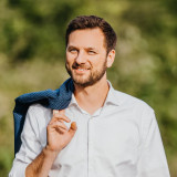 Daniel Staiger im Vordergrund, weißes Hemd mit hochgekrempelten Ärmeln und das Jacket über die Schulter geworfen. Im Hintergrund eine gründe Landschaft aus Hecke und Rasen. 