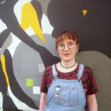 Eine weiße, junge Frau mit Brille und rotbrauner Ponyfrisur steht in einer Latzhose vor einer mit Graffiti verzierten Wand