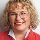 Tatjana Bautsch Portraitfoto einer blonden, lächelnden Frau mit welligem, mittellangem Haar, schmal eingefasster großer Brille, die eine weiße Bluse und einen knallroten Mantel trägt.