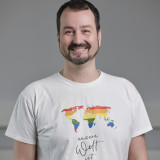 Mann mittleren Alters mit Bart. Schwarze Haare, lächelnd. Weißes T-Shirt mit Regenbogenfarben darauf. Arme hängen runter.