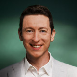 Daniel Belling Eine Portraitaufnahme des Kandidaten Doktor Daniel Belling. Er trägt ein helles Sakko mit weißem Hemd und schaut gerade und mit einem lächelnden Gesichtsausdruck in die Kamera. Der Hintergrund ist dunkelgrün.