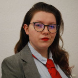 Zu sehen ist eine junge, aufstrebende Politikerin. Sie trägt einen grauen Blazer, ein hellblaues Hemd und eine rote Krawatte. 