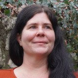 Eine dunkelhaarige Frau steht lächelnd vor einer efeubewachsenen Mauer. Es ist nur das Gesicht zu sehen.