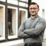 Selmar Ibrahimović Junger Mann Mitte 20. Er trägt eine braune, halbrunde Brille und einen Seitenscheitel mit braunen Haaren. Er lächelt freundlich und hält die Arme vor dem Körper kämpferisch verschränkt. Er trägt ein graues Oberteil mit Kragen und steht vor einem Fachwerkhaus mit hell ausgeleuchteten Fenstern.