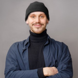 Leon Turi Kley Junger, freundlicher Mann. Schwarze Beanie, Schwarzer Rollkragenpullover darüber eine langärmliges dunkelblaues Hemd. Oberlippenbart mit 3-Tage-Bart, Ohrringe auf beiden Seiten und einem seitlichen Nasenpiercing. Posiert in Selbstbewusster Haltung, mit verschränkten Armen vor dem Körper.