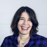 Katja Bühring schaut offen und mit einem Lachen den Betrachter an. Sie trägt ein lila-schwarz kariertes Karohemd und eine Kette mit einem kleinen Fisch als Anhänger.