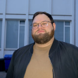 Porträt eines Mannes mit Brille und Bart, der draußen in einem Hinterhof steht. Er trägt eine schwarze Jacke über einem braunen Pullover und blickt mit neutralem Gesichtsausdruck in die Kamera.