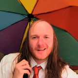 Das Bild zeigt einen jungen Mann mit langen Haaren und Halbglatze. Er trägt ein weißes Hemd mit einer schlecht gebundenen roten DIE PARTEI-Krawatte. Er lächelt etwas verpeilt und hat einen Regenbogenschirm in der Hand.