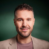 Porträt einer erwachsenen männlich gelesenen Person mitte Dreißig mit hellem Teint. Er hat kurze, leicht gelockte dunkelbraune Haare mit kürzeren Seiten, braune Augen und einen kurzen Bart. Er blickt direkt in die Kamera und lächelt leicht. Er trägt ein beigefarbenes Sakko über einem roséfarbenen T-Shirt. Der Hintergrund ist einfarbig dunkelgrün und unscharf, das Licht ist weich und gleichmäßig.