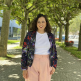 In der Mitte des Bilds steht eine junge Frau mit lockigen braunen Haaren. Sie trägt eine rosa Hose, ein weißes T-shirt und eine blumige Jacke. Im Hintergrund sind Bäume zu sehen. 