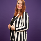 Isabelle Krause Das Bild zeigt die Kandidatin von der Hüfte aufwärts vor einem lilanen Hintergrund mit einem schwarz weiß gestreiften Blazer.