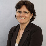 Esther Kanschat Ein sehr freundlich, lächelndes Gesicht, braune kurze Haare, braunes Jacket mit beidem Top darunter. Randlose Brille, arme vorm Körper verschränkt, aber locker