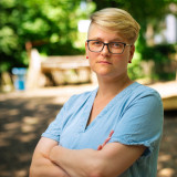 Das Bild zeigt Maria Stockhaus mit verschränkten Armen und ernstem Blick. Maria Stockhaus trägt ein hellblaues Jeanskleid, eine Brille, kurze blonde Haare und rote Ohrstecker. Der Hintergrund ist von Grün geprägt und leicht verschwommen.