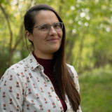 Luise Globig Porträt von Luise Globig mit Brille und langen braunen Haaren vor einem grünen, unscharfen Waldhintergrund. Sie trägt eine weiße Bluse mit kleinen Fuchsmotiven.