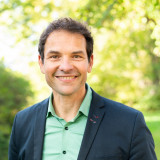 Martin Stümpfig Das Bild zeigt Martin Stümpfig, einen Mann mit kurzen dunklen Haaren, der lächelt. Martin Stümpfig trägt ein grünes Hemd und einen dunkelblauen Blazer