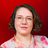 Morgan Gwendolyn Varbelow Porträt einer Frau vor kräftig rotem Hintergrund. Sie blickt frontal in die Kamera und lächelt leicht. Sie trägt eine runde Metallbrille, einen kleinen Nasenring und eine bunt gemusterte Bluse in Rosa-, Orange- und Weißtönen. Ihre dunkelblonden, kinnlangen Haare sind seitlich gescheitelt und leicht gewellt. Die Haltung wirkt aufrecht und ruhig, der Gesichtsausdruck freundlich und selbstbewusst.