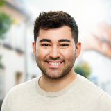 Das Bild zeigt einen jungen Mann mit dunklem, lockigem Haar und einem vollen Bart. Er lächelt freundlich in die Kamera und trägt einen hellbeigen Pullover. Der Hintergrund ist unscharf und zeigt eine Außenumgebung mit Bäumen und Gebäuden.