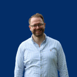 Ruven Gastel Ruven Gastel vor einem dunkelblauen Hintergrund. Er trägt ein hellblau-weiß gestreiftes Hemd und eine Brille. Er lächelt.