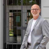 Ralf Krings im grauen Anzug und weißem T-Shirt auf den Stufen vor dem Krefelder Rathaus. Linke Hand in der Hosentasche. Ralf Krings lächelt, trägt eine Brille. Er hat einen kahlen Kopf und einen weißen 3-Tage-Bart.