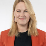 Weiße Frau mit mittellangen blonden Haaren in den Dreißigern mit einen organenen Blaser und schwarzem Oberteil.Sie lächelt und hat die Haare nach vorne, aber auf einer Seite sind die Haare hinter das Ohr gesteckt mit einem großen goldenen Ohrring.