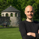 Der Kandidat steht mit verschränkten Armen auf einer grünen Wiese. Im Hintergrund ist ein runder Turm aus Kalkstein zu sehen.