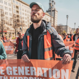 Theodor Schnarr nimmt an einem sonnigen Tag an einem Protestmarsch der Bewegung Letzte Generation teil. Er trägt eine orangene Warnweste und hält ein Banner, auf dem steht "Letzte Generation vor den Kipppunkten". Um ihn herum sind weitere Menschen zu sehen. Er lächelt leicht und schaut fokussiert nach vorne.