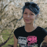 Foto, Porträt, halber Oberkörper. Freundlich lächelnde Frau mit zurückgebundenem, blauem Haar; im Hintergrund ist, leicht unscharf, ein blühender Kirschbaum zu sehen.