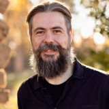 Tobias Kremkau Porträt von Tobias Kremkau mit zurückgekämmten dunklem Haar und langem Vollbart. Er trägt ein schwarzes Oberteil und blickt direkt in die Kamera; im unscharfen Hintergrund ist eine herbstlich beleuchtete Parklandschaft zu sehen.