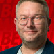 Marco Rumza Alternativtext für das Bild: Porträt eines Mannes mit Brille und kurzem, blond-grauem Haar, der freundlich und selbstbewusst in die Kamera blickt. Er trägt ein dunkles Sakko über einem hellen T-Shirt. Im Hintergrund ist eine rote Fläche mit grafischen SPD-Schriftzügen zu sehen.