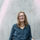 Susanne Jahn vor einer grauen Wand. Lange Haare, Brille, dunkelgrünes Hemd, bunte Kette