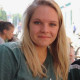 Sie sehen eine junge Frau Mitte 20, grünes T-Shirt, blonde mittellange Haare, freundlich lächelnd, im Hintergrund ein Cafe