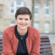 Sara Nanni, in einer roten Jacke und schwarzem Oberteil, sitzt auf einer Bank in Düsseldorf und lächelt in die Kamera.