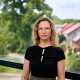 Andrea Leonhard Es ist eine Frau mit blonden Haaren und Brille auf dem Foto zu sehen. Sie trägt ein schwarzen T-Shirt und eine dunkelgrüne Hose. Im Hintergrund sind verschwommen Böume und Hausdächer zu erkennen.