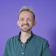 Mathew Bristow Ein Profilbild von Volt-Kandidat Matt Bristow. Er steht vor einem lila Hintergrund und schaut lächelnd in die Kamera.
