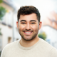 Das Bild zeigt einen jungen Mann mit dunklem, lockigem Haar und einem vollen Bart. Er lächelt freundlich in die Kamera und trägt einen hellbeigen Pullover. Der Hintergrund ist unscharf und zeigt eine Außenumgebung mit Bäumen und Gebäuden.