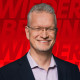 Stefan Schenck Weißer Mann mit Brille und grau-meliertem Haar. Im dunklen Sakko mit fliederfarbenem Hemd. Lächelt vor einem roten Hintergrund in die Kamera.