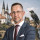 Daniel Wald im dunkelblauen Anzug mit weißem Hemd und bordeauxfarbener Krawatte. Im Hintergrund sind der Merseburger Dom und Schloss, sowie eine Rabenstatue aus der Rabensage, mit dem Ring zu sehen.