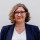 Julia Schlembach Bild einer Frau mit kinnlangen gelockten Haaren und Brille. Sie trägt ein weißes Shirt und einen dunkelblauen Blazer.