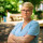 Maria Stockhaus Das Bild zeigt Maria Stockhaus mit verschränkten Armen und ernstem Blick. Maria Stockhaus trägt ein hellblaues Jeanskleid, eine Brille, kurze blonde Haare und rote Ohrstecker. Der Hintergrund ist von Grün geprägt und leicht verschwommen.