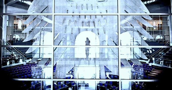 Blick durch die Glasfassade in den Plenarsaal des Deutschen Bundestags. Im Vordergrund sind die großen, stilisierten Bundesadler-Elemente auf der Glaswand zu sehen. Dahinter erkennt man die blauen Sitzreihen des Plenarsaals, Treppen und einzelne Personen. Im Zentrum steht silhouettenhaft eine Person vor einem hell erleuchteten Durchgang. Das Bild ist kontrastreich und in kühlen Blau- und Grautönen gehalten.